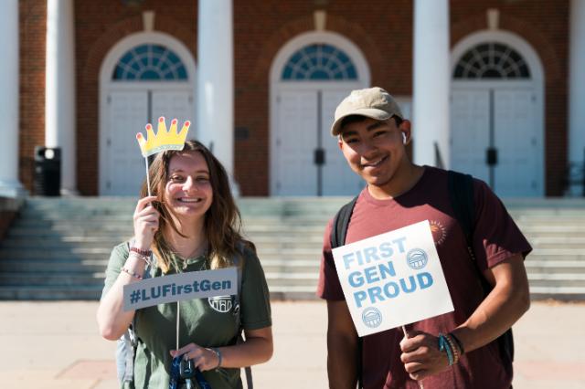 - Two students smile while holding signs celebrating first-generation college pride. One holds a crown prop and a sign reading “#LUFirstGen.” The other holds a sign reading “First Gen Proud.” They stand in front of a campus building with columns in bright daylight.