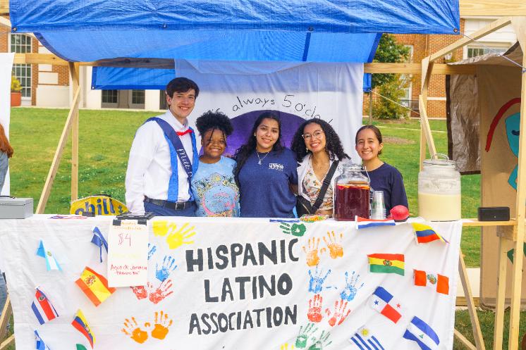Hispanic Latino Association at Oktoberfest - Five students stand behind a booth for the Hispanic Latino Association. A banner with colorful handprints and flags decorates the front of the table. They smile at the camera with drinks and supplies displayed. The booth is set up outdoors on campus with buildings in the background.