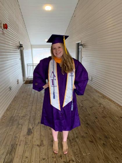 Alisa Kish in academic regalia.  - A graduate stands smiling in a hallway wearing a purple cap and gown. She poses with hands on her hips while wearing honor cords and a stole. The stole shows Greek letters and a Class of 2019 designation. The wooden walkway and white walls create a simple corridor setting.