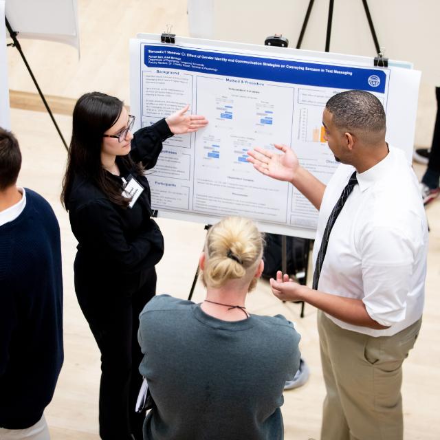 - Student presents a research poster while discussing the project with attendees. The presenter gestures toward charts and text sections on the academic poster. Two listeners stand nearby and engage in conversation about the research. The scene takes place during a busy poster session with other displays nearby.