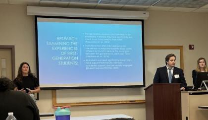 - Three students present research in a classroom using a projected slide titled “Research Examining the Experiences of First-Generation Students.” One student stands at a podium while another operates a computer beside the screen. A third student stands to the side holding notes during the presentation. Desks, a projector screen, and audience members are visible in the classroom setting.