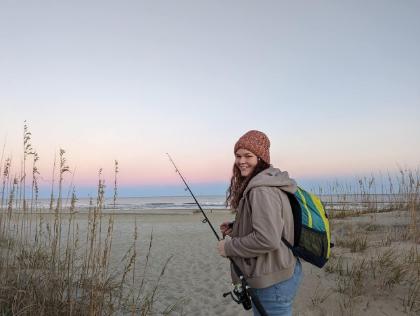 - A student stands on a sandy beach holding a fishing rod at sunset. She wears a knit hat, jacket, and backpack while smiling toward the camera. Sea oats and dunes frame a path leading to the shoreline. Soft pink and blue evening colors stretch across the sky over the ocean.