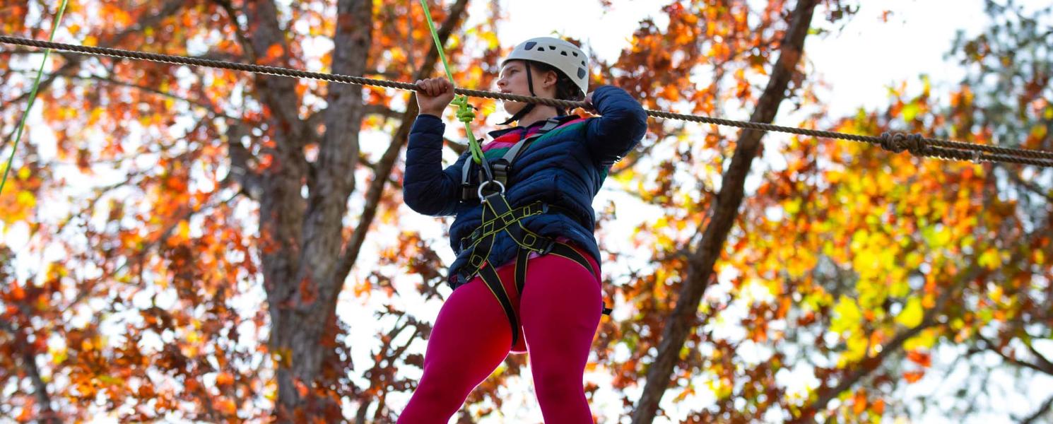 A student wearing a helmet and harness crosses a ropes course among fall trees.