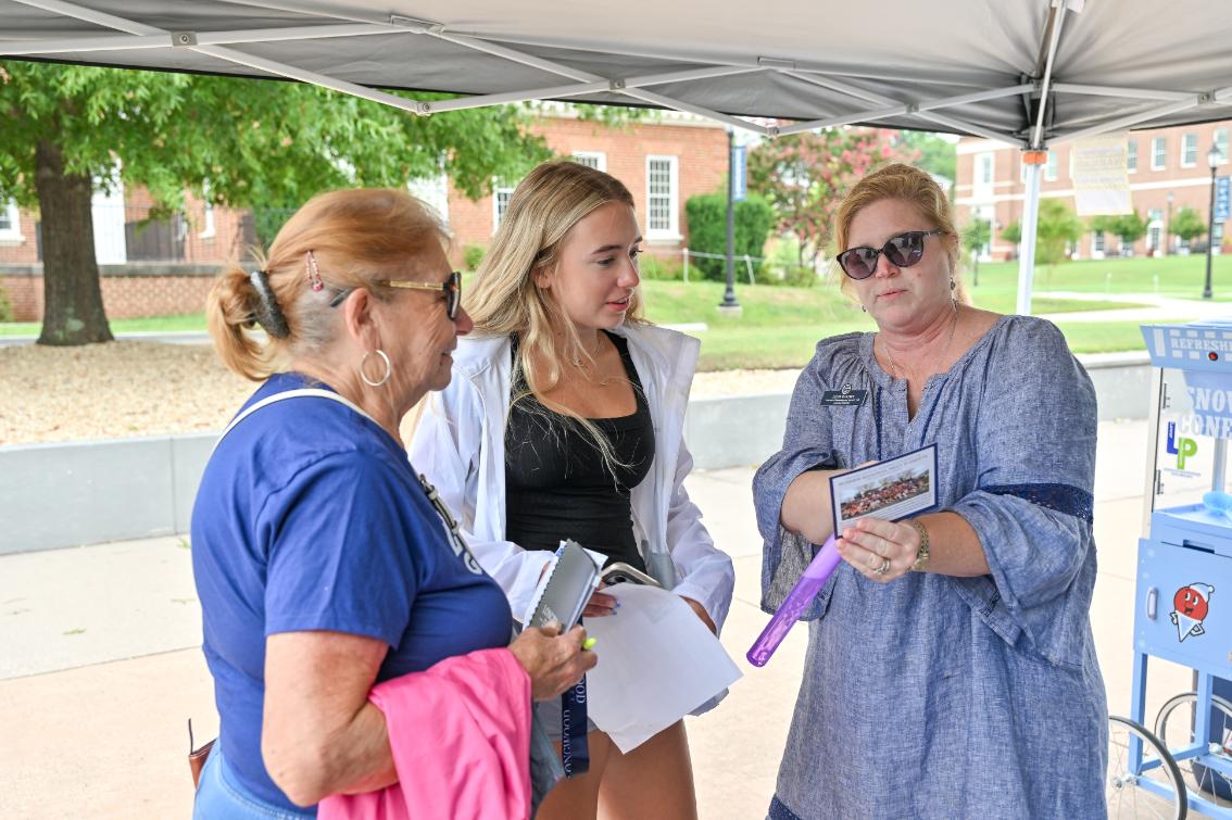 Family gets help during an orientation session - Staff member speaks with two visitors under a tent on campus. She holds a card and gestures while explaining information. The visitors listen while holding papers and a notebook. Conversation takes place at an outdoor orientation information table.