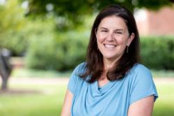 Maria Hamilton - Woman with long brown hair smiles while standing outdoors on campus. She wears a light blue top and simple jewelry. Trees and greenery blur softly in the background. Portrait-style photo taken in natural daylight.