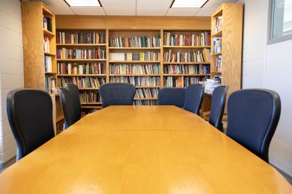 Chairs, Table, and stand full of books - A long wooden conference table sits in the center of a small meeting room. Black office chairs surround the table on both sides. Tall wooden bookshelves filled with books line the back wall. Fluorescent ceiling lights illuminate the quiet study and meeting space.