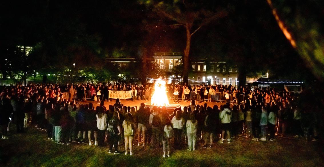 CHI Burning at Longwood University - Large crowd gathers at night around a bonfire on a campus lawn. Flames rise from a fire pit as students stand in a wide circle. Metal barricades line the area with trees and string lights in the background. A campus building glows behind the crowd in the dark.
