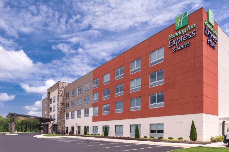 Holiday Inn Express - Farmville - Exterior of the Holiday Inn Express & Suites building under a bright, partly cloudy sky. The four-story hotel has red and tan panels with rows of windows. A covered entrance canopy extends toward an empty parking lot. Holiday Inn Express & Suites signs are mounted near the roofline.
