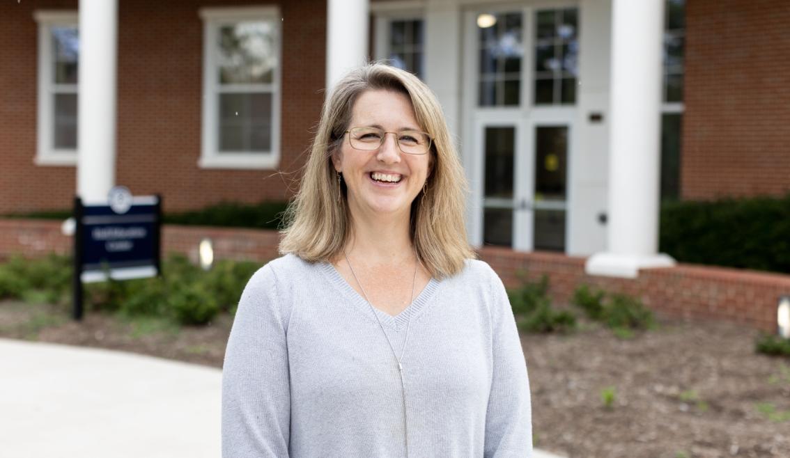 Angela McDonald - Woman with shoulder-length blonde hair and glasses smiling outside a brick campus building. She wears a light gray sweater and a long silver necklace. White columns and glass doors frame the entrance behind her. Landscaping and a campus sign are visible in the softly blurred background.