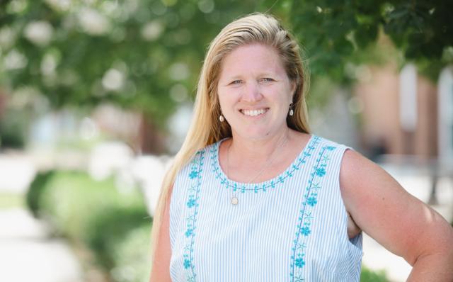 Julie Wiley Ramsey ’93 - Woman with long blonde hair smiling outdoors beneath leafy green trees. She wears a sleeveless blue-and-white striped blouse with turquoise embroidery. A silver necklace and drop earrings complete her look. Softly blurred campus buildings and greenery appear in the background.