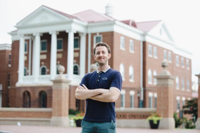 Dr. Jay Crowell ‘09 - Man standing with arms crossed in front of a red brick campus building with white columns. He wears a navy polo shirt and green pants. The historic building features tall columns and arched windows. Brick pillars and landscaping frame the foreground.