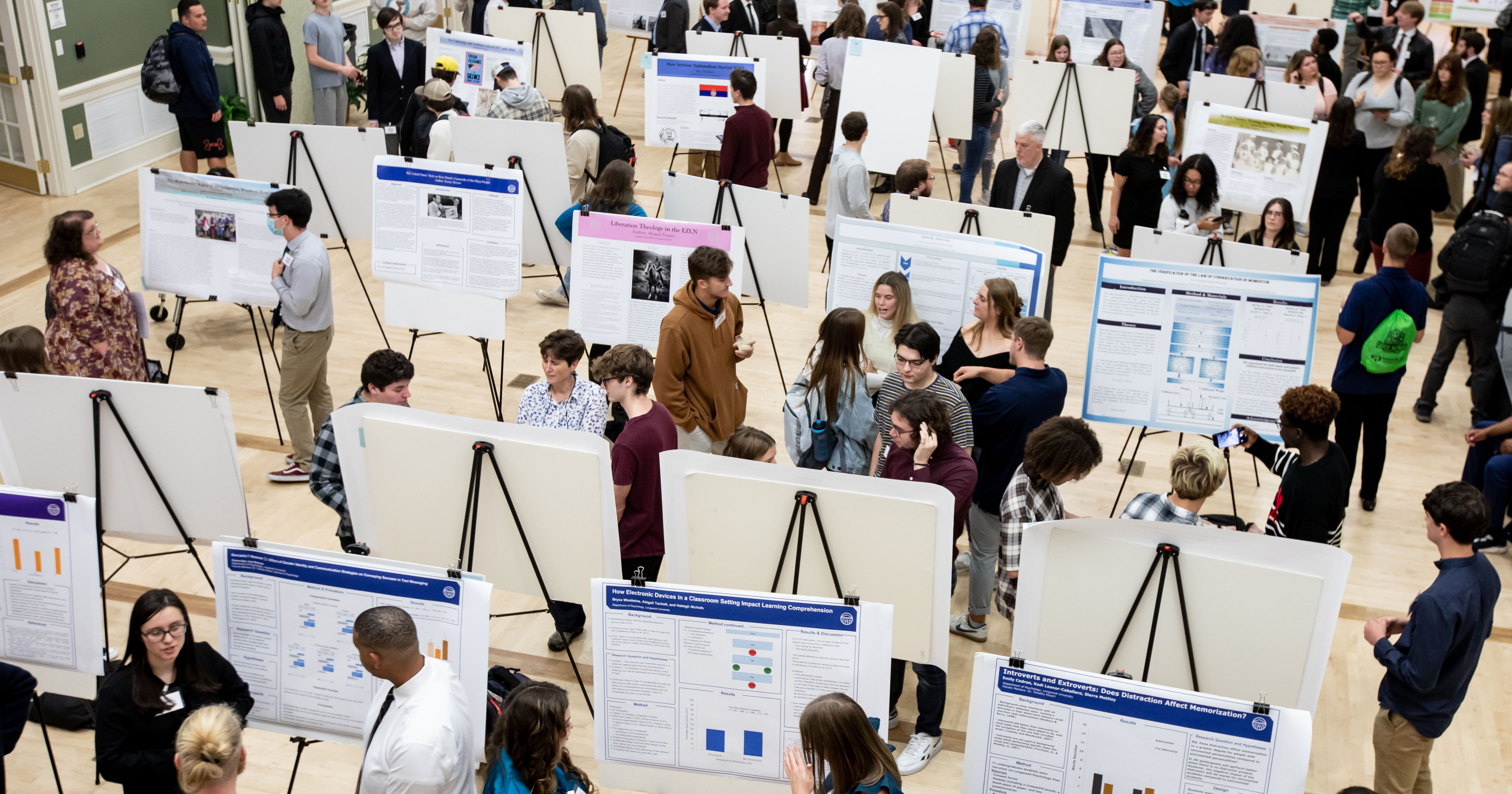 Research Day - Crowd of students and faculty gathered around research posters in a large hall. Poster boards on easels fill the space in neat rows across the floor. Small groups discuss findings, pointing to charts and printed data. The busy showcase highlights a campus-wide academic research event.