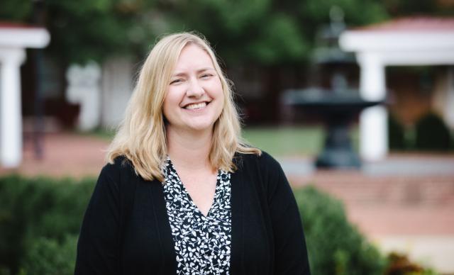 Sara Neher - Woman with shoulder-length blonde hair smiling outdoors on campus. She wears a black cardigan over a black-and-white patterned blouse. A brick courtyard and decorative fountain appear behind her. Greenery and white architectural columns frame the background.
