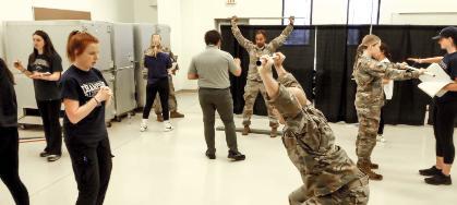 Kinesiology students screening soldiers - Students observe National Guard members during a hands-on training session. One soldier performs a deep overhead stretch with a weighted bar. Others conduct mobility and strength assessments in small groups. The activity takes place in a large indoor classroom space.
