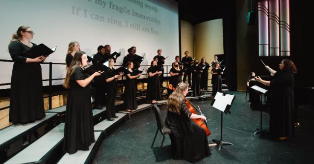 Choir rehearsing in Jarman - A choir performs on stage dressed in formal black attire. Singers stand on tiered risers holding sheet music. A conductor leads the ensemble with raised arms. A cellist plays in front of the group during the performance.