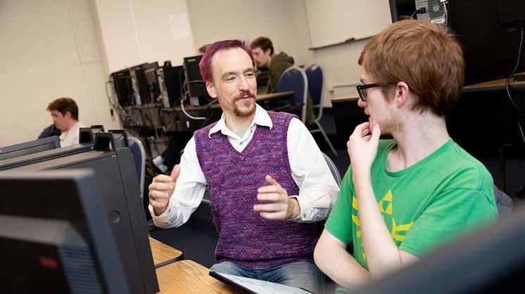 Don Blaheta with a student - A professor gestures while speaking with a student at a computer workstation. They sit side by side in a computer lab filled with desktop monitors. The student listens thoughtfully during the discussion. Other students work at computers in the background.