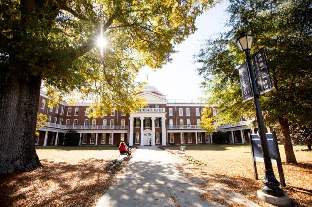 Rotunda with sun peeking through the trees and two students sitting on a bench out front