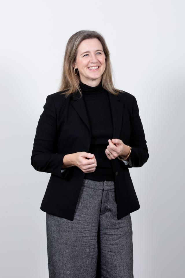 Dr. Angela McDonald - A woman with shoulder-length light brown hair stands against a light background. She wears a black blazer over a black turtleneck with gray trousers. She smiles while gesturing lightly with her hands. The portrait is framed from mid-thigh up.