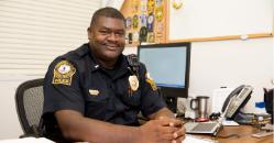 Lieutenant John Johnson - A Longwood University police officer sits at a desk in his office. He wears a dark uniform with a badge and shoulder patches visible. A computer monitor, phone, and laptop sit on the desk beside him. He looks at the camera with a friendly, professional expression.