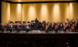Richmond Symphony in Jarman Auditorium - A full orchestra performs on a concert hall stage beneath warm overhead lights. The conductor stands on a podium at center, leading the ensemble. Musicians play strings, brass, woodwinds, and harp behind music stands. Audience members sit in shadow in the foreground.