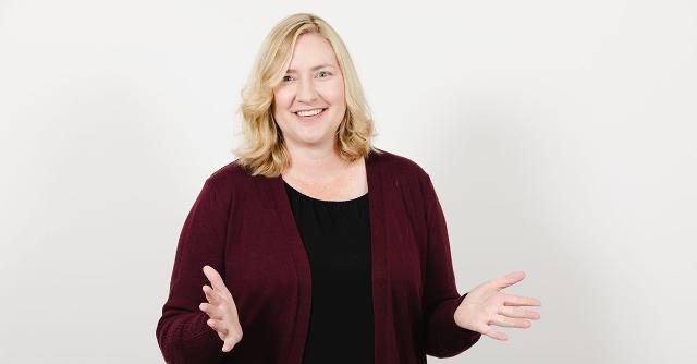 Sara Neher - A woman with shoulder-length blonde hair stands against a light background. She wears a black top with a maroon cardigan. Her hands are open as if speaking or presenting. She smiles warmly at the camera.