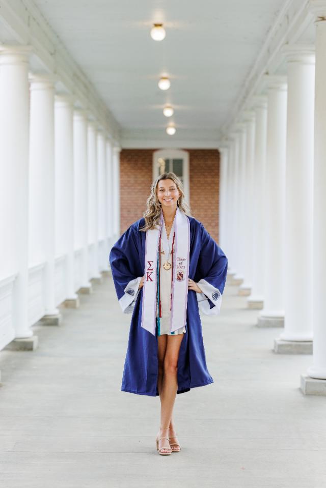Madison Poe ’23 - A graduate in a navy gown walks confidently through a white columned colonnade. She wears a stole with Greek letters and “Class of 2023” embroidered on it. Honor cords and a medal hang around her neck. The portrait captures a poised graduation moment on campus.