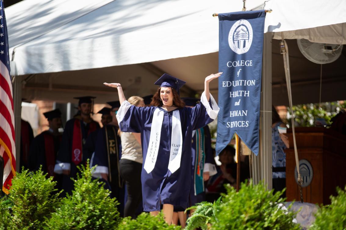 Graduate student wearing a first generation sash holds her hands in the air as she crosses the stage