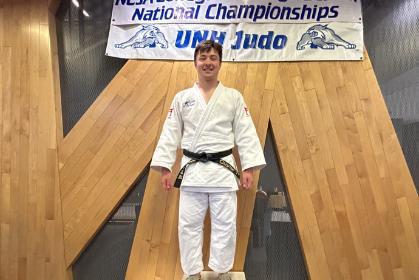 Kosei Cuyler ’26 - Judo athlete in a white gi and black belt stands on the first-place podium. A banner above reads “NCJA Collegiate & High School National Championships” and “UMW Judo.” The three-step podium shows numbers 2, 1, and 3. Wood paneling and gym flooring frame the awards area.