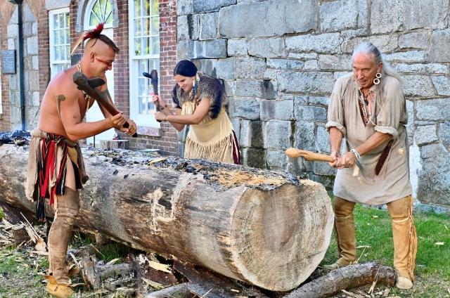 Russell Reed (on left) cutting into a log with swings of his axe, recreating the Native American art form of constructing canoes.