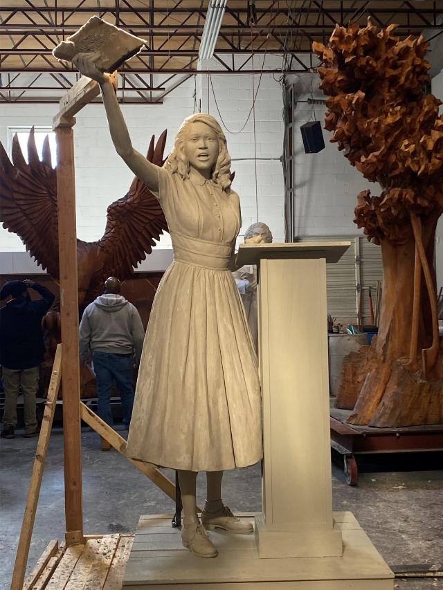 The Barbara Johns model. Courtesy of Virginia Department of Historic Resources. - A full-size sculpted figure of Barbara Johns stands in an indoor workshop. She raises an arm holding papers beside a simple podium. Large carved props, including wings and a tree, surround the statue. Workers and scaffolding appear in the background under exposed rafters.