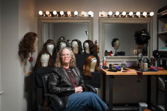 Samantha Wootten '00 sits in a dressing room chair surrounded by mannequin heads with wigs. She wears glasses, a black leather jacket, and long pants, with hands folded in her lap. Lighted mirrors and styling tools line the workspace around her. Multiple wigs in various styles and colors are arranged behind and beside her.