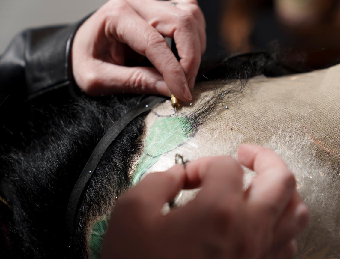 Samantha Wootten's hands use a small tool to attach strands of hair to a wig on a mannequin head. Individual hairs are being carefully threaded into a mesh base. Pins and marked sections guide the precise placement of the hair. The close-up highlights the detailed craftsmanship of wig making.