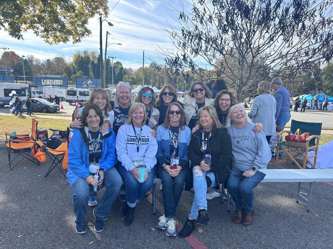 As an alumna and two-time Longwood parent, Kathy Hansen Fox '85 (back row, second from left), has long made Farmville a getaway destination with friends, family and fellow Lancers. - Group of Longwood alumni poses together at an outdoor tailgate event. Many wear Longwood apparel and lanyards, smiling in front of tents and chairs. A Longwood banner and vehicles appear in the background near athleti