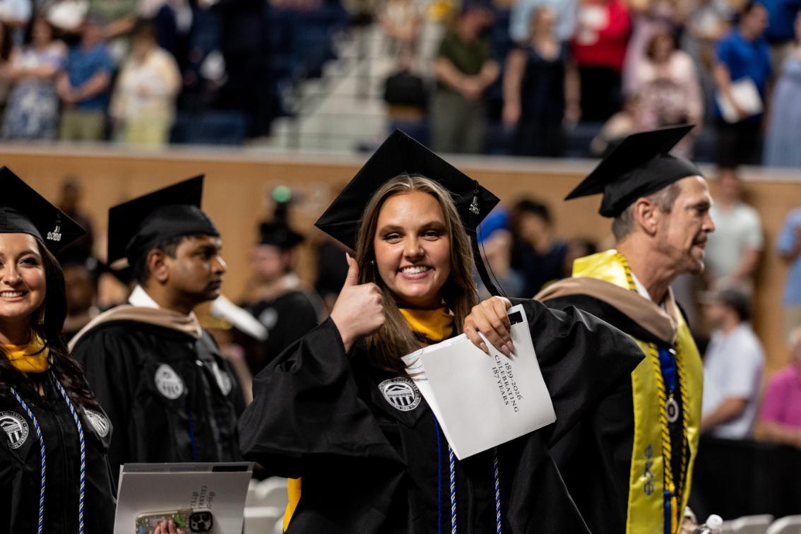 A smiling Longwood University graduate in a black cap and gown gives a thumbs-up while holding a Commencement program during the 2026 Graduate Commencement ceremony.