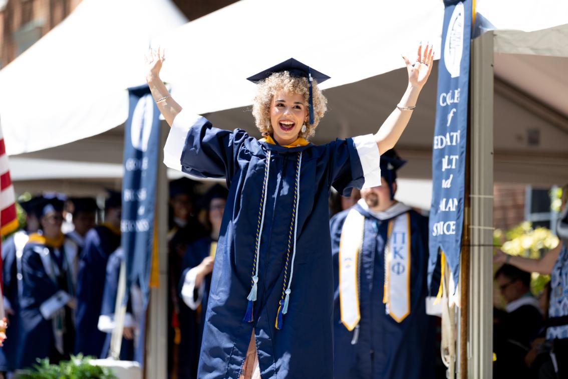 A Longwood University undergraduate graduate in a blue cap and gown smiles with both arms raised while walking across the stage during Commencement.