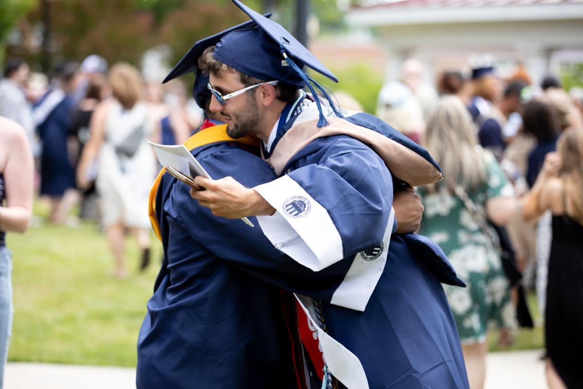 A Longwood University undergraduate graduate embraces a classmate after the Commencement ceremony, surrounded by family and friends on campus.