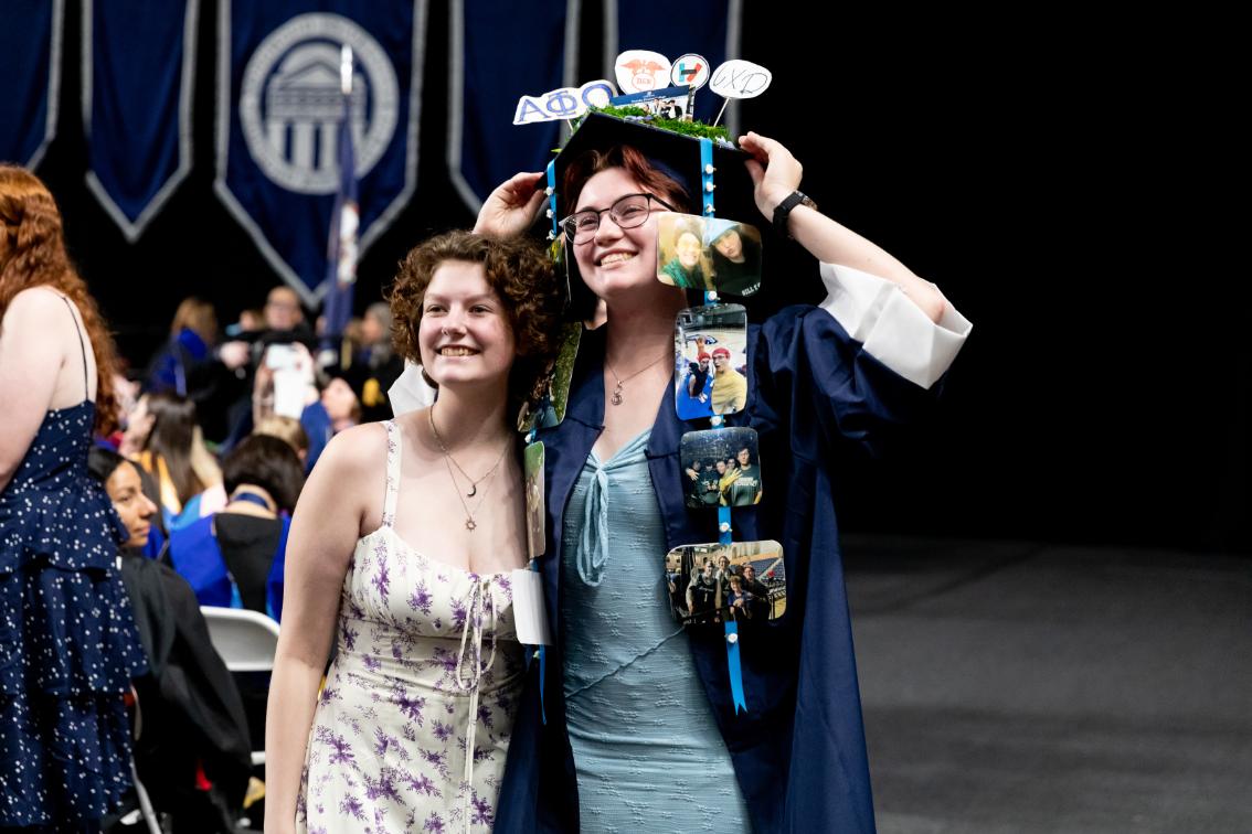 Graduate in a navy cap and gown smiles with a friend during Convocation, wearing a decorated mortarboard with photos and mementos.