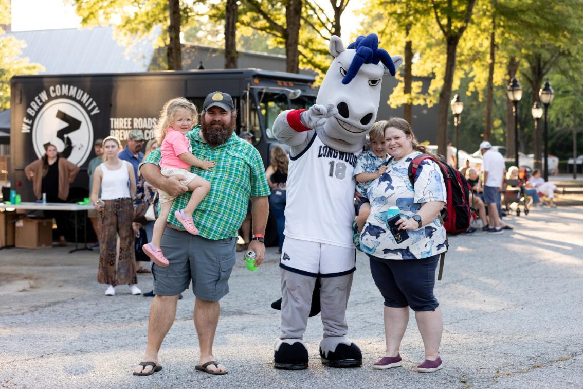 Elwood poses with a smiling family during Longwood Family Weekend, with a food truck and campus guests in the background.